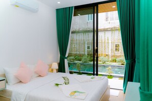 A bedroom with a bed, pillows, a nightstand, a lamp, and a view of a swimming pool and greenery outside.