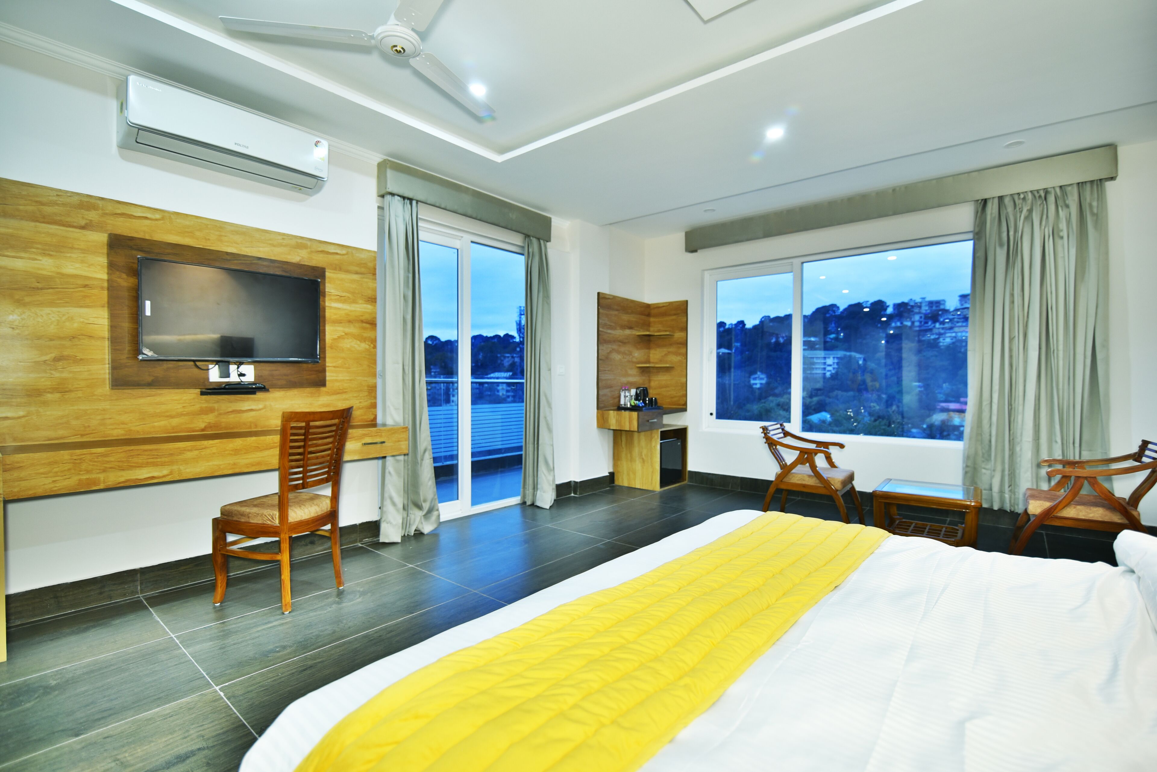 Habitación de hotel con una cama grande, un televisor montado en la pared, un escritorio con silla y vistas a la ciudad a través de amplios ventanales.