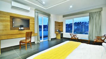 Habitación de hotel con una cama grande, un televisor montado en la pared, un escritorio con silla y vistas a la ciudad a través de amplios ventanales.