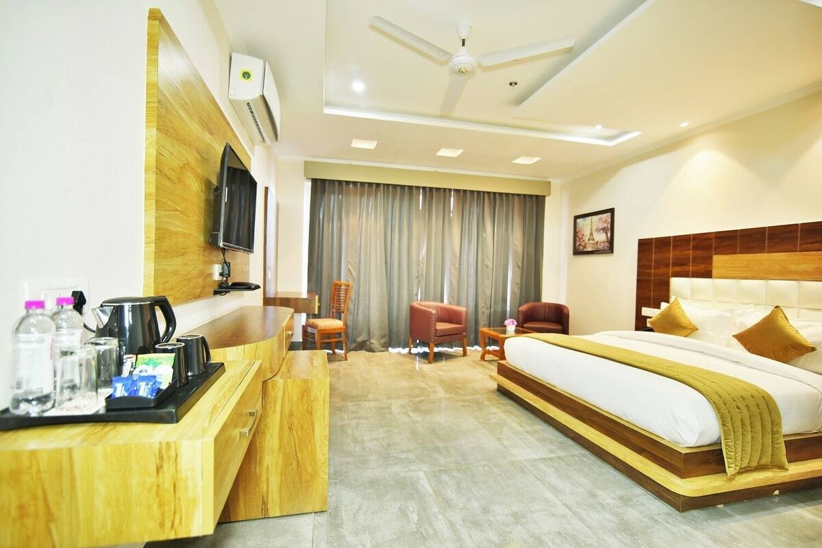 A hotel room with a bed, a desk with a coffee maker, a TV, a chair, a sofa, and a window with curtains.