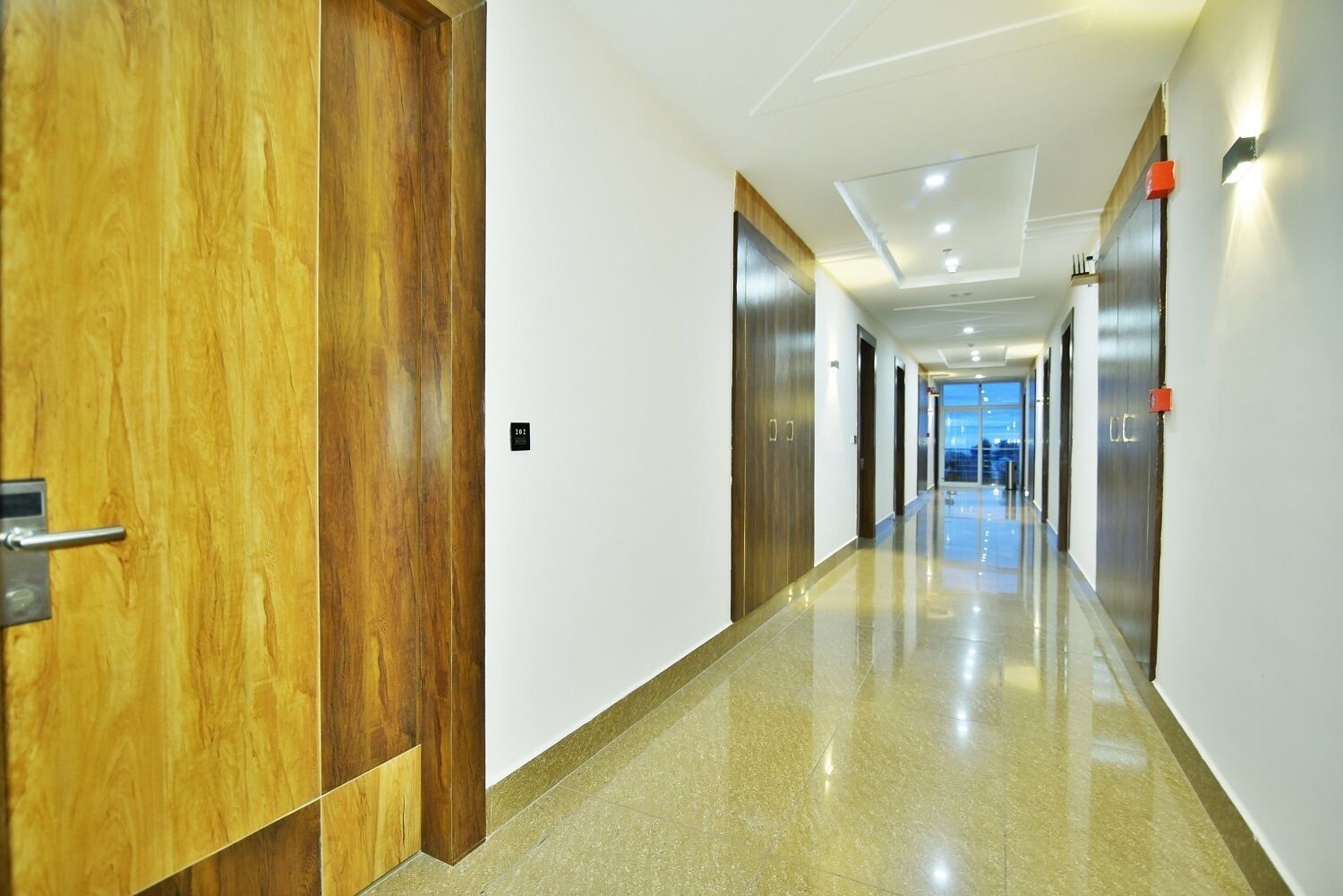 A modern hotel corridor with wooden doors, a shiny floor, and a view of the city at the end.