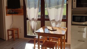 Ein Zimmer mit Holztisch und -stühlen, einem Fernseher auf einem Regal, einer Mikrowelle und Blick auf Grünfläche durch das Fenster.