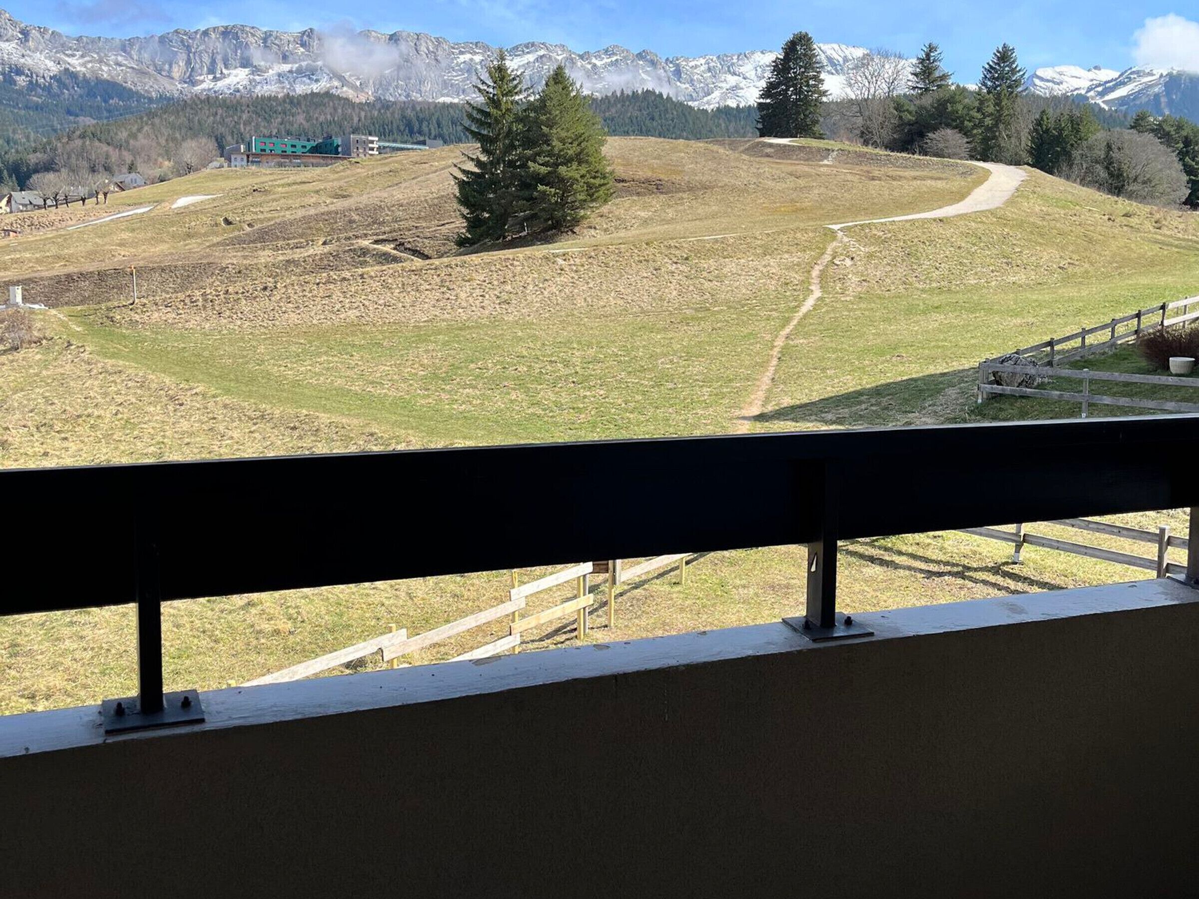 Une vue depuis un balcon donnant sur un paysage vallonné, avec un sentier, une clôture en bois et un bâtiment au loin.