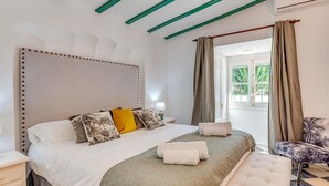 Quarto com uma cama grande, um banquinho, uma cadeira e vista para o exterior.
