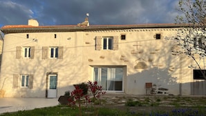 A two-story building with a tiled roof, a large sliding glass door, and a small garden with red and blue flowers. - Charming Farmhouse Retreat: Farm Life, Wildlife & Scenic Views, Perfect for Family Holidays (Autrac)