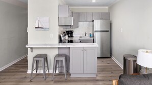 A compact kitchen with grey cabinets, a white marble countertop, and a stainless steel refrigerator. There are two stools and a small trash bin. - Sunshine Paradise: Oceanfront Retreat Awaits (North Topsail Beach)