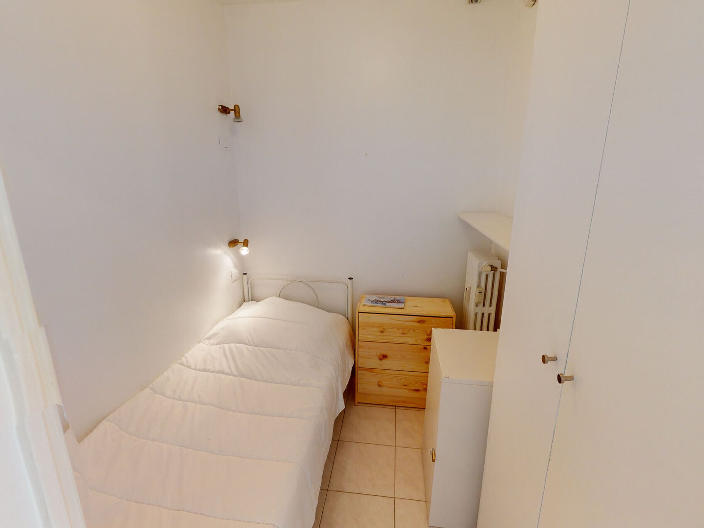 A small, compact bedroom with a single bed, a wooden dresser, a radiator, and a wardrobe.