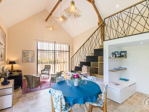 A spacious living area with a high ceiling, a dining table with chairs, a staircase with a geometric railing, and a large window with vertical blinds.