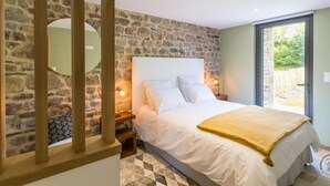 Une chambre avec un mur en pierre, un lit recouvert de draps blancs, une tête de lit en bois, une table de chevet avec une lampe, et une vue à travers une porte vitrée.