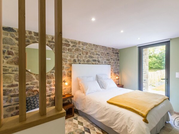 A bedroom with a stone wall, a bed with white linens, a wooden headboard, a nightstand with a lamp, and a view through a glass door. - Comfortable house with private garden in Réville (Réville)