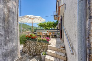 Escadaria de pedra que leva a um terraço com área de estar ao ar livre e uma mesa, tudo cercado por vasos de plantas e um grande guarda-sol.