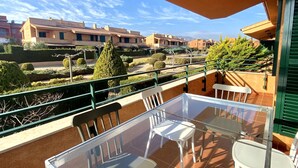 Un balcon avec une table et des chaises en verre, donnant sur un quartier résidentiel où les haies sont soigneusement taillées et où le ciel est d’un bleu éclatant.