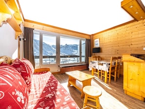A cozy room with a wooden interior, a sofa, a dining table, and a view of snow-covered mountains through large windows.