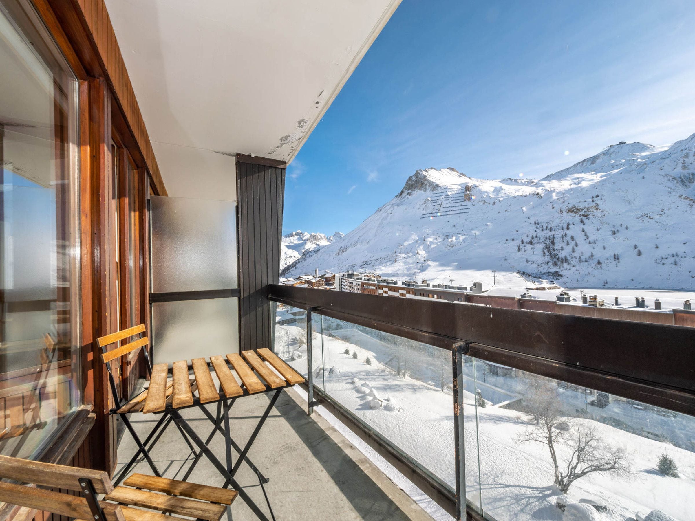 Un balcón con sillas de madera, un ventanal de vidrio y vistas a montañas nevadas.
