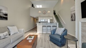 Una sala moderna con un sofá gris, un sillón azul y una mesa de centro de madera. Una escalera conduce a un nivel superior con una zona de cocina.