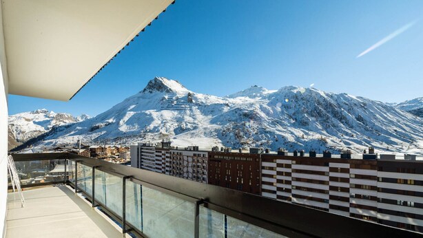 Ein Balkon mit Glasgeländer, der Blick auf einen schneebedeckten Berg und Gebäude freigibt.