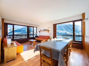A spacious room with a wooden table and chairs, a sofa, and a balcony with a mountain view.
