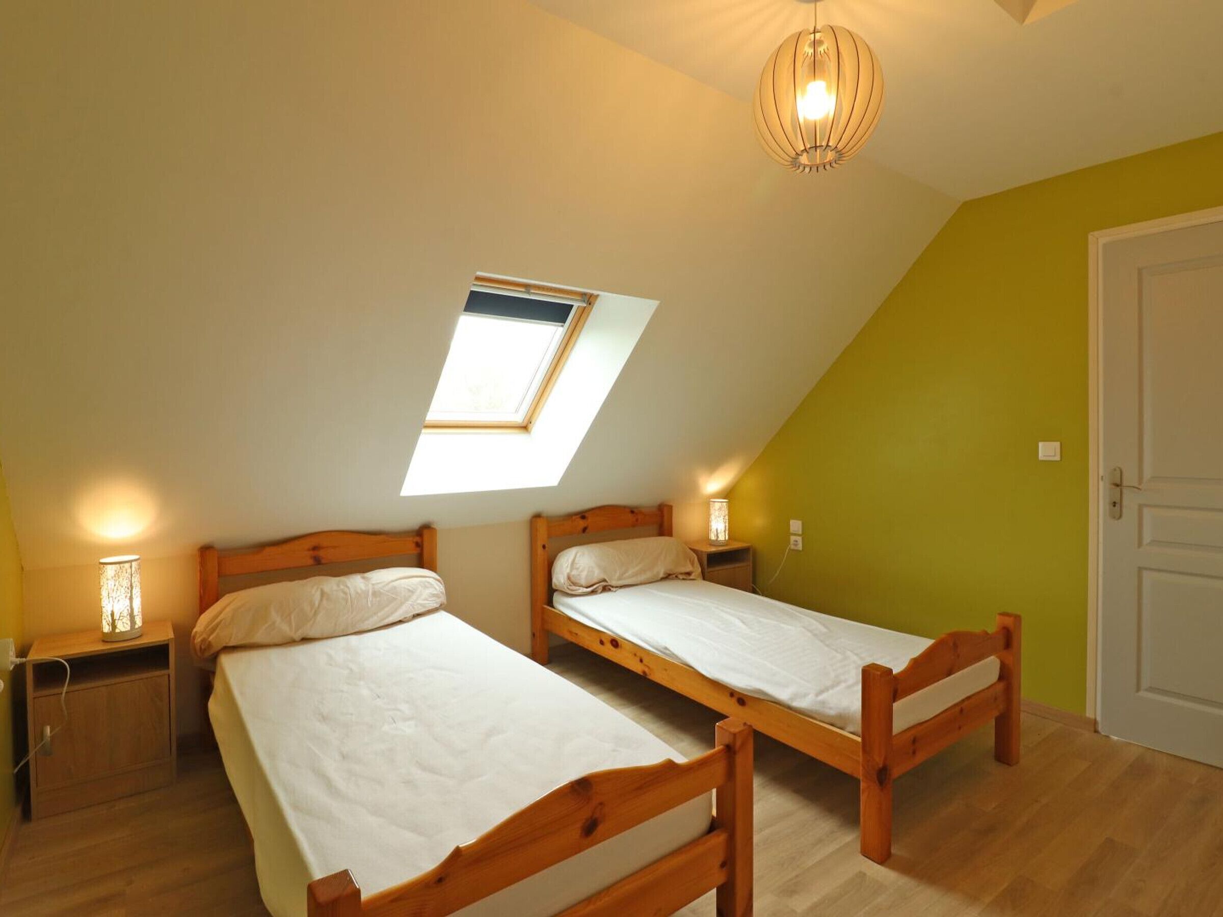 A small, cozy room with two wooden beds, a skylight, a hanging light fixture, and a door.