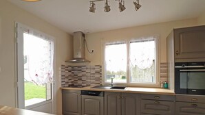 A kitchen with a wooden table, a built-in oven, and a window with lace curtains. - Renovated house with enclosed garden, 3 bedrooms, free EV charging, near Swiss Normandy (Sainte-Opportune)