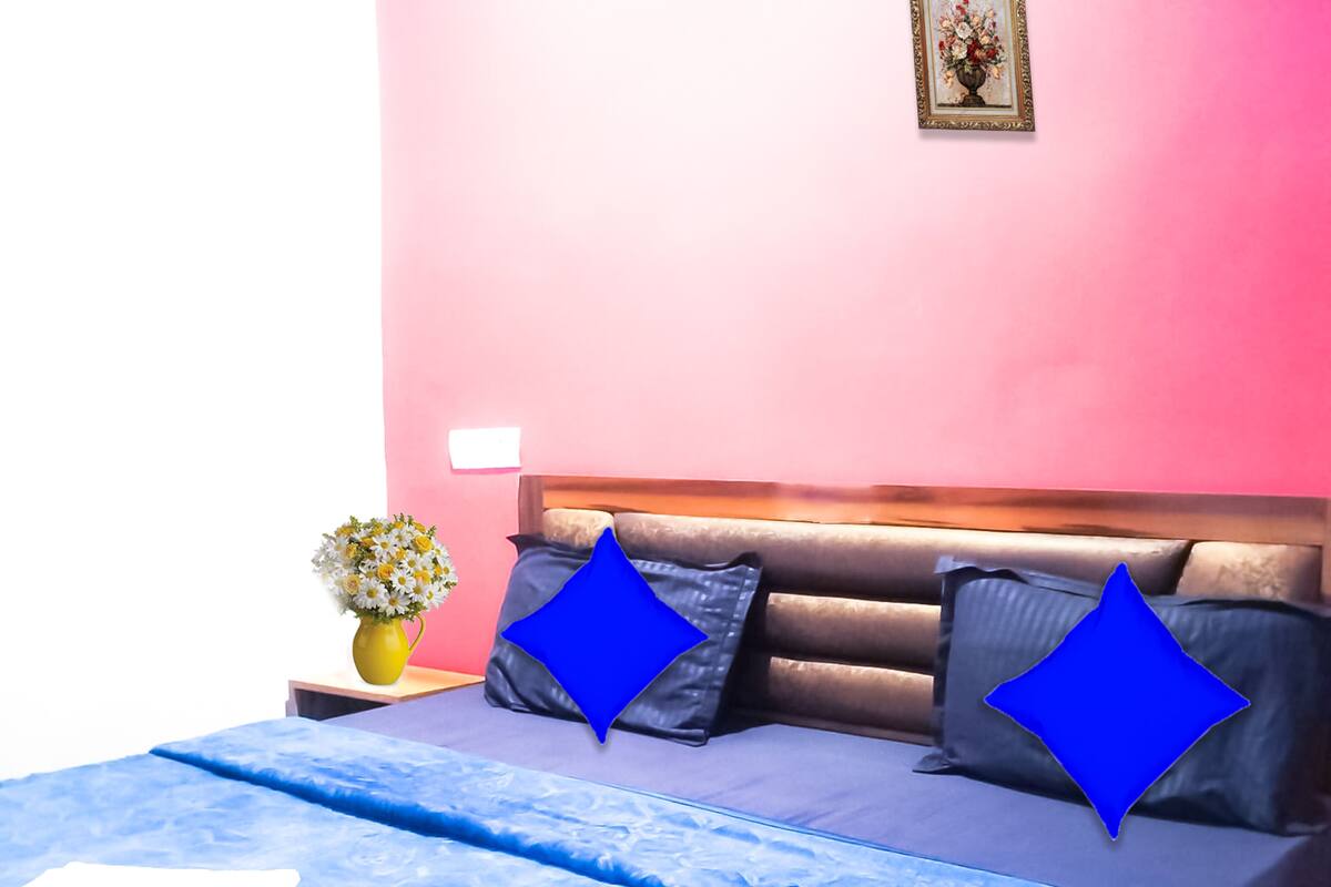 A bed with a blue bedspread and pillows, a yellow vase with flowers, and a pink wall.