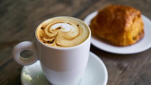 A white cup of coffee with latte art on a saucer, placed on a wooden table. - Roomshala 021 Hotel Joyous Inn (Ghaziabad)