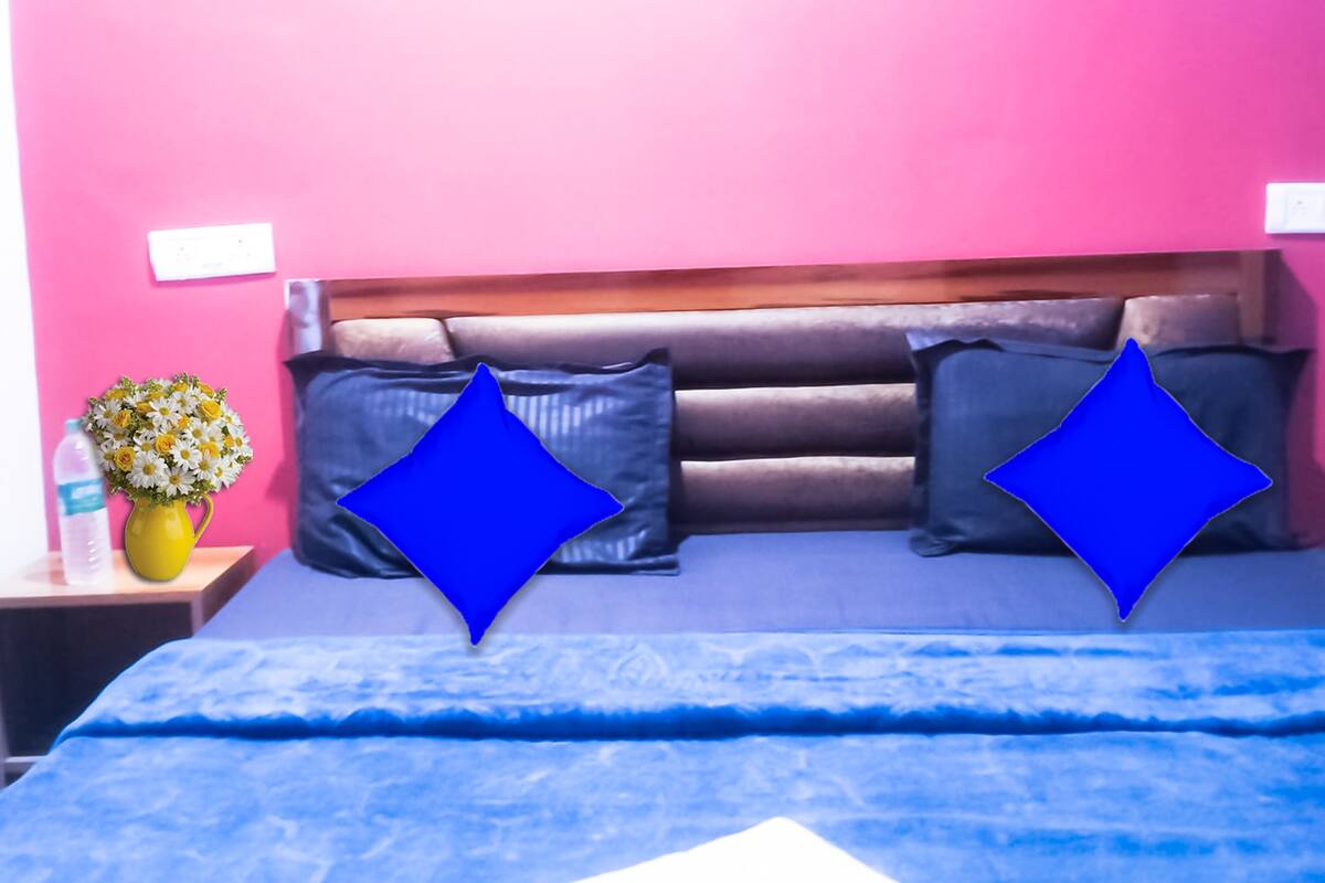 A bed with a blue bedspread, two pillows, a wooden headboard, and a nightstand with a water bottle and a vase of flowers.