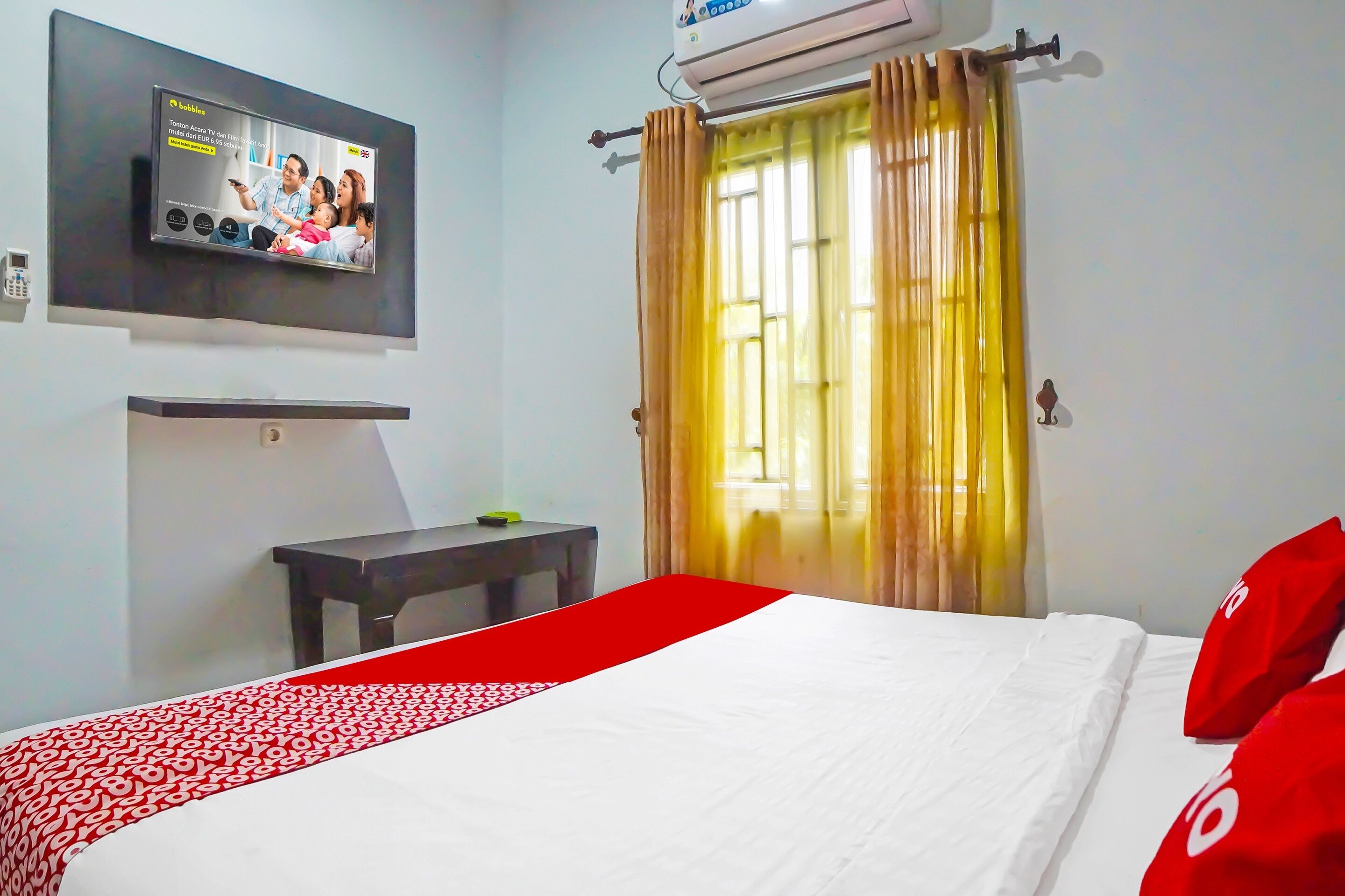 A hotel room with a bed, a TV mounted on the wall, a small table, and a window with curtains.