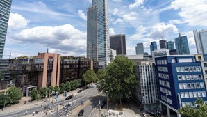 Un paisaje urbano con edificios modernos altos, cielo despejado y una calle concurrida con peatones y vehículos.