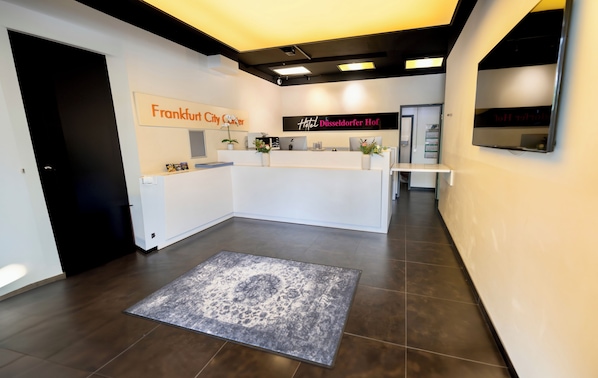 A hotel reception area with a modern design, featuring a reception desk, a large rug, and a sign indicating the hotel's name.