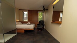 Una habitación de hotel moderna con una cama, una silla, un espejo grande y una ventana que ofrece vistas a edificios y árboles.