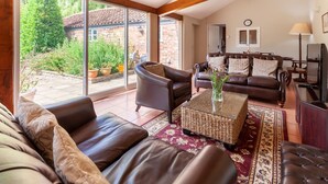 A spacious living room with leather sofas, a coffee table, and a view of a garden through large windows. - The Cow Barn - sleeps 6 guests  in 3 bedrooms (Banningham)
