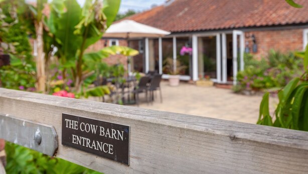 A rustic barn entrance with a sign, surrounded by lush greenery and a patio area with outdoor seating. - The Cow Barn - sleeps 6 guests  in 3 bedrooms (Banningham)
