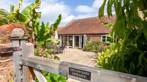 A rustic barn entrance with a wooden gate, surrounded by lush greenery and a patio area. - The Cow Barn - sleeps 6 guests  in 3 bedrooms (Banningham)