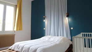 A bedroom with a bed, a crib, a window with yellow curtains, and a wall-mounted light fixture.