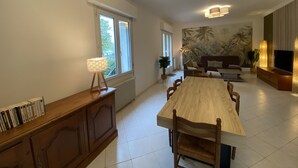 A spacious living room with a large wooden dining table, a sofa, and a TV. There are windows providing natural light and a mural on the wall.