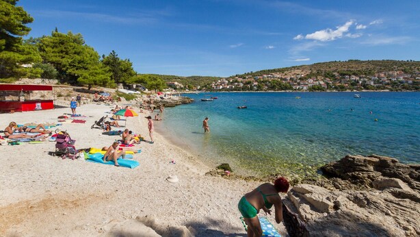 Une plage à l’eau claire, où l’on peut voir des gens se prélasser au soleil et des bateaux au loin.