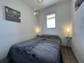 Een slaapkamer met een bed, twee bedlampen, een raam met zonwering, een wandairconditioner en een lijst met een afbeelding.
