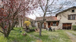 A rustic stone house with a garden, a small outdoor seating area, and a visible staircase leading to the upper floor. - 65 sqm cottage with fireplace, terrace, and children's play area (Saint-Médard-en-Forez)