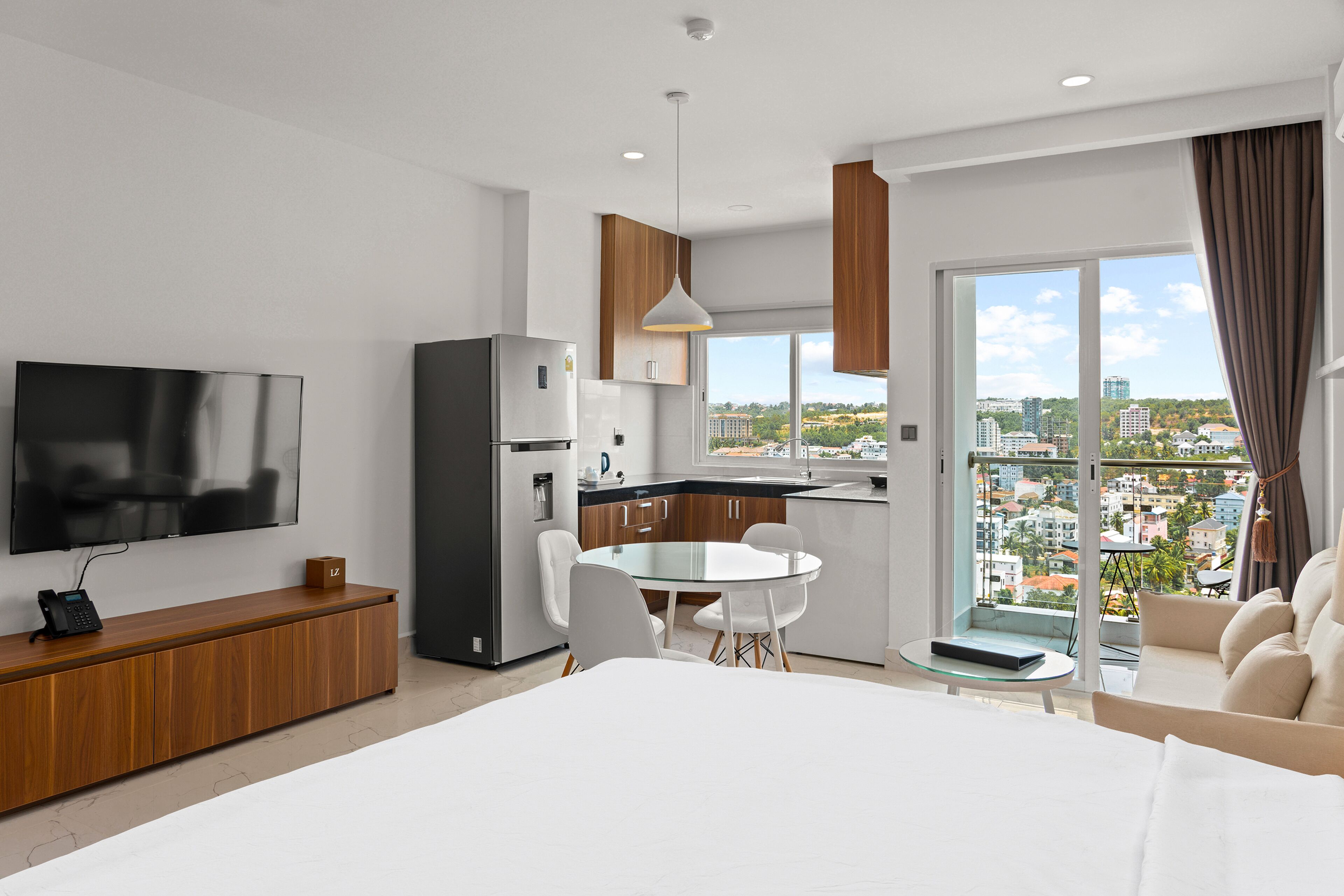 Une chambre d’hôtel moderne dotée d’un grand lit, d’un coin repas avec une table en verre, d’une cuisine équipée d’un réfrigérateur et d’un téléviseur fixé au mur.
