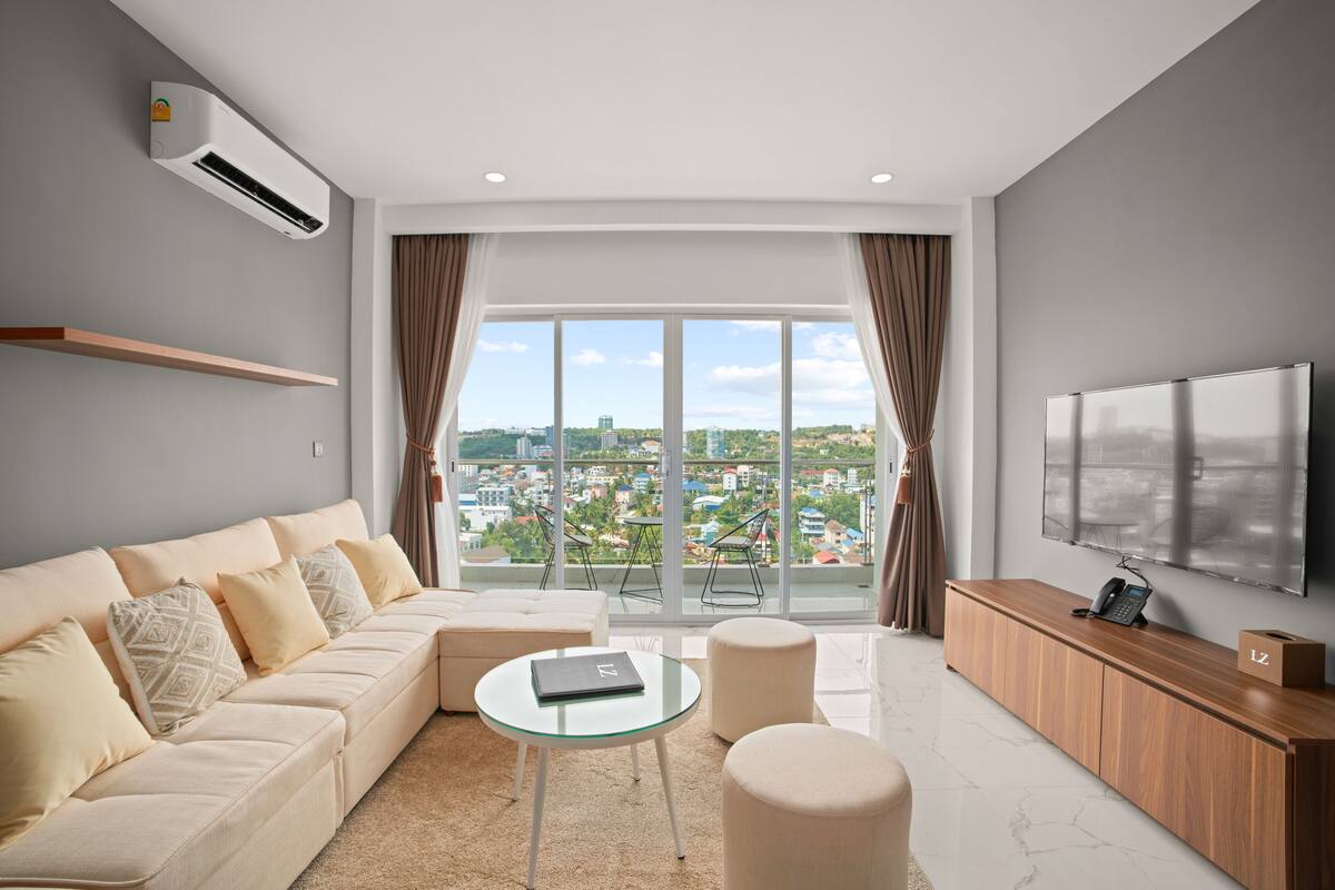 A modern living room with a sofa, glass coffee table, ottoman, and a flat-screen TV.