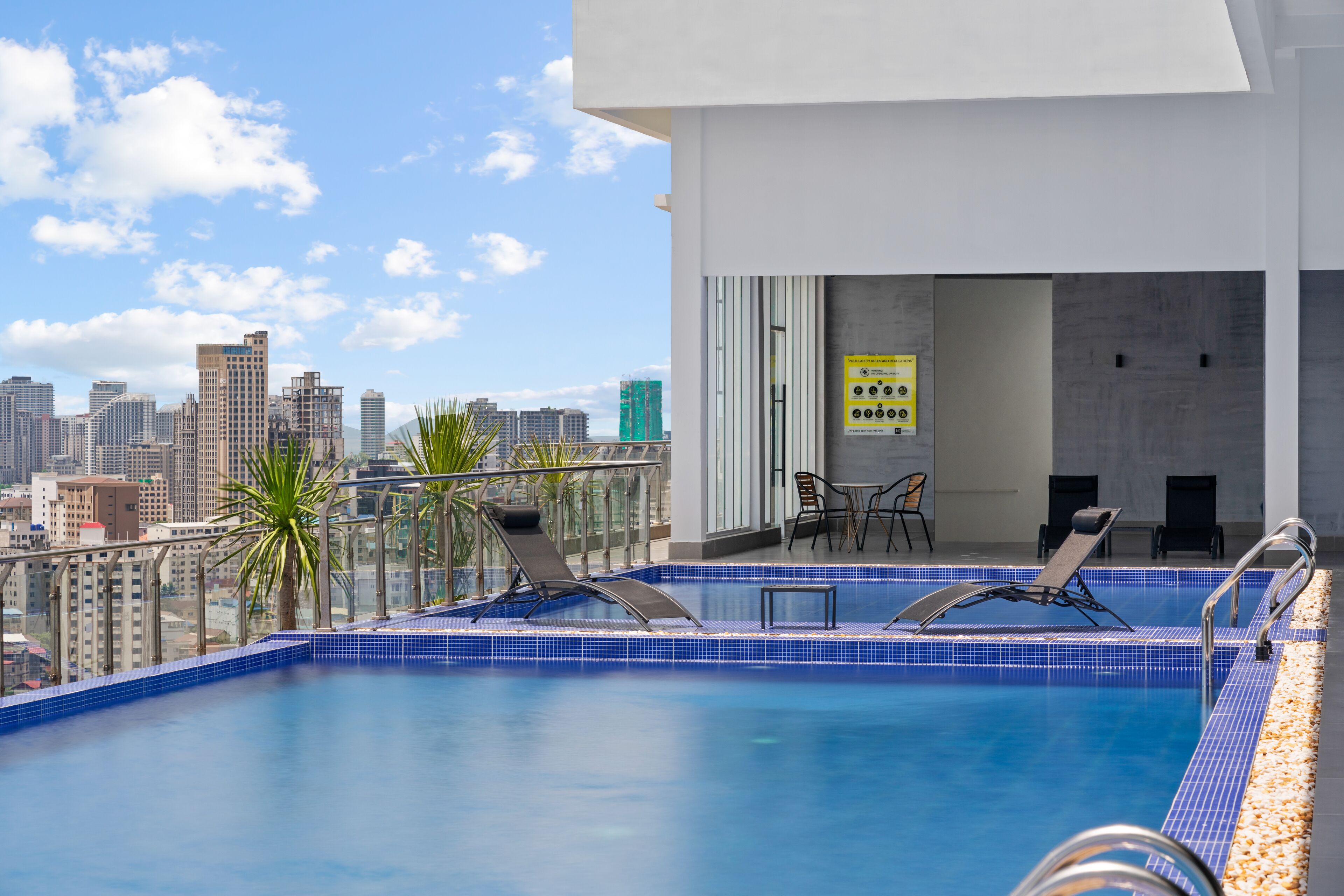 A rooftop pool with two sun loungers, a small table, and a chair. A city skyline is visible in the background.