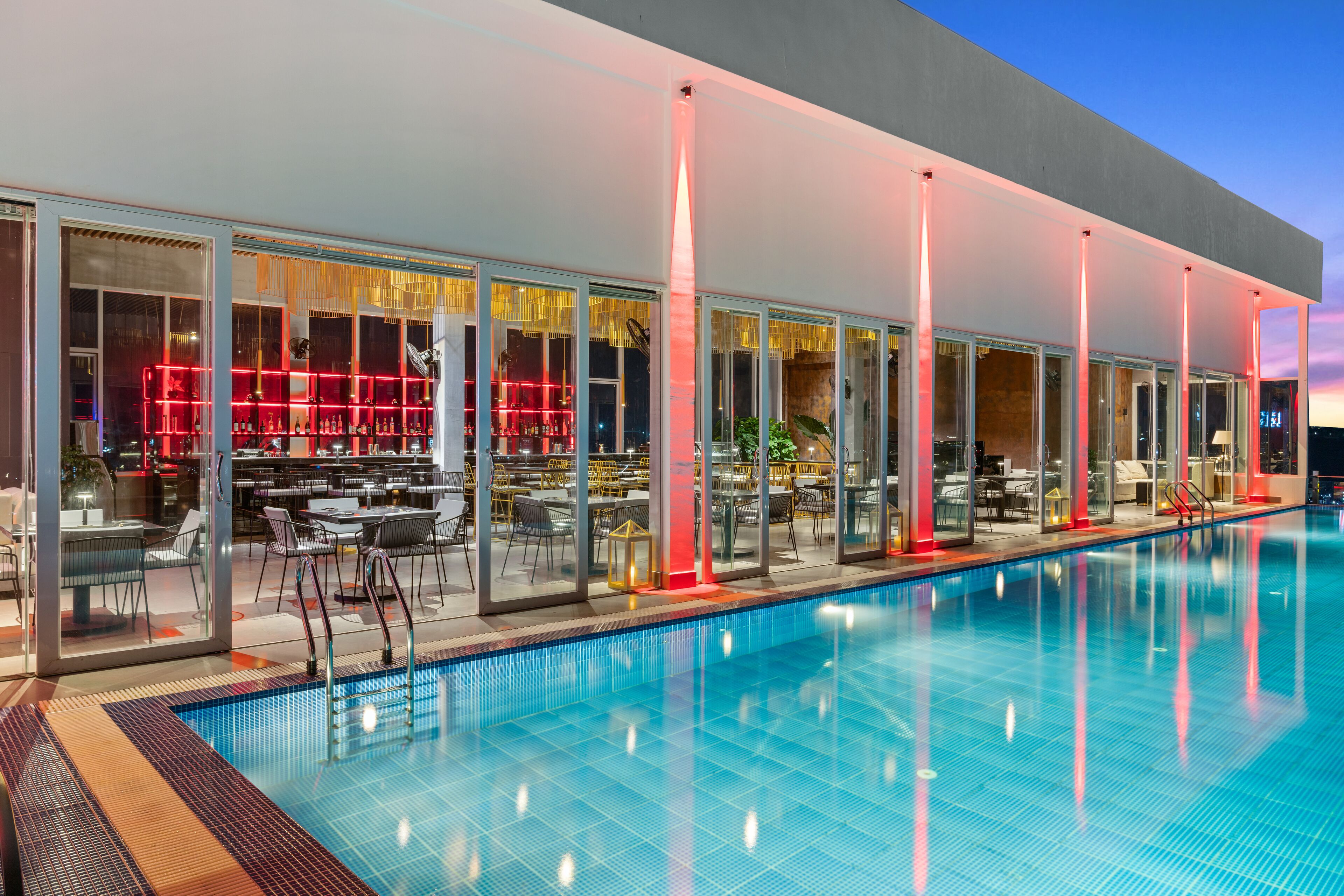 A modern hotel with a swimming pool, glass-enclosed dining areas, and red ambient lighting.