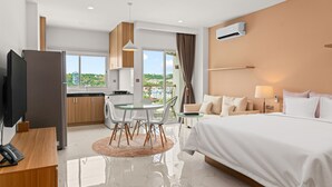 A modern hotel room with a large bed, a desk with a phone, a dining area with chairs, and a view of the city. - MLZ APARTMENTS (Sihanoukville)
