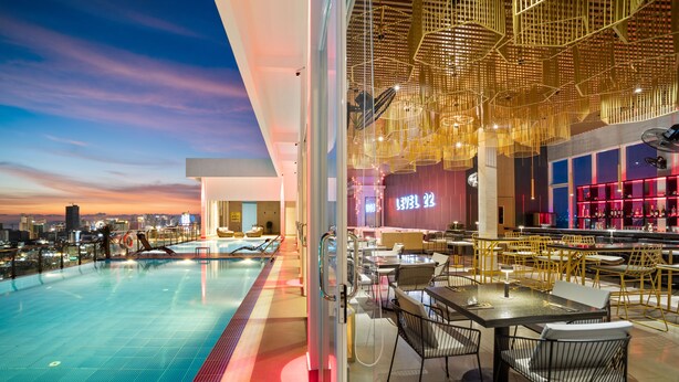 A modern indoor pool area with a city view, a bar with neon signage, and a dining area with tables and chairs. - MLZ APARTMENTS (Sihanoukville)