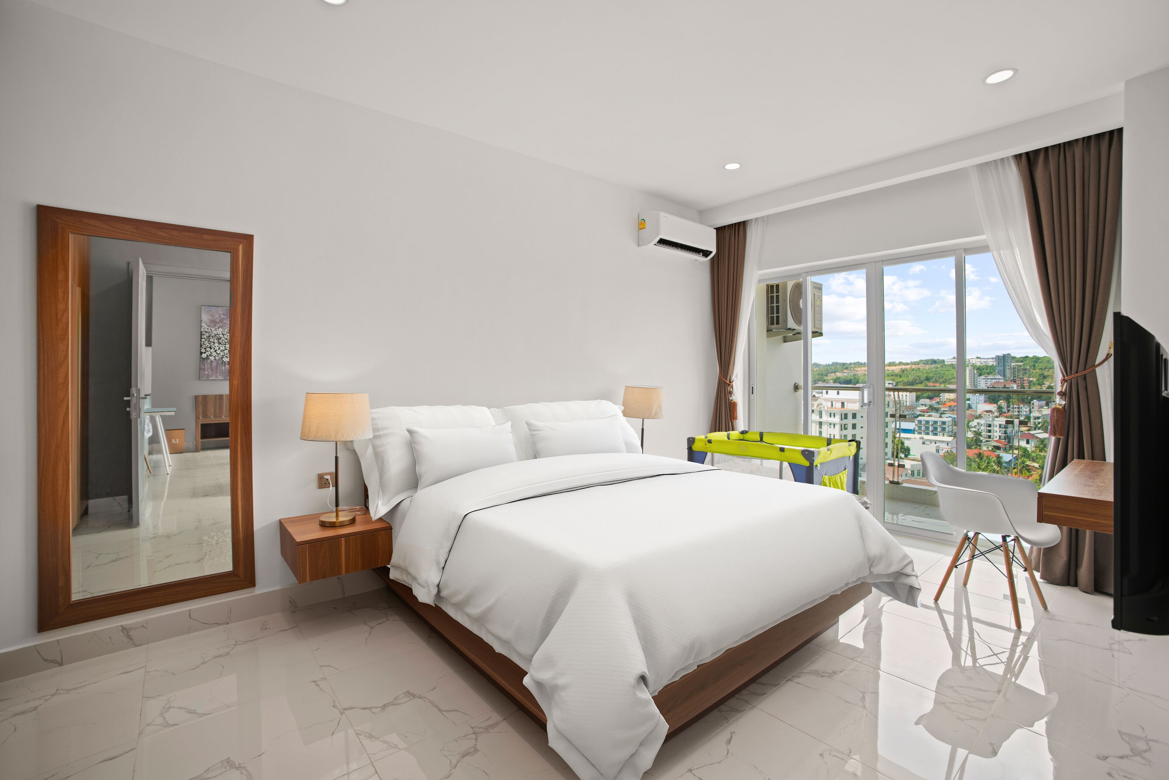 Habitación de hotel moderna con una cama grande, un espejo, una mesita de noche con lámpara, vistas a la ciudad a través de amplios ventanales y un balcón con una silla.