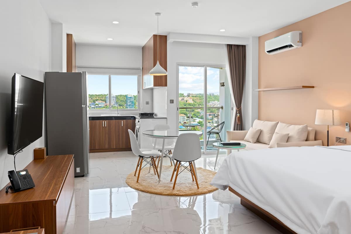 A hotel room with a bed, a desk with a TV, a dining area with chairs, a kitchen, and a balcony with a view of buildings.