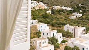 A view of a coastal village with white buildings, a prominent hill in the background, and a window with white blinds.