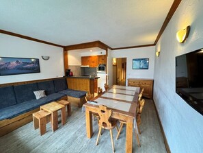 Un espace de vie compact comprenant un canapé, une table à manger, des chaises en bois, un coin cuisine et un téléviseur fixé au mur.