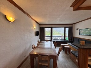 A dining area with a long table and benches, a television mounted on the wall, and a view of a mountainous landscape through large windows.