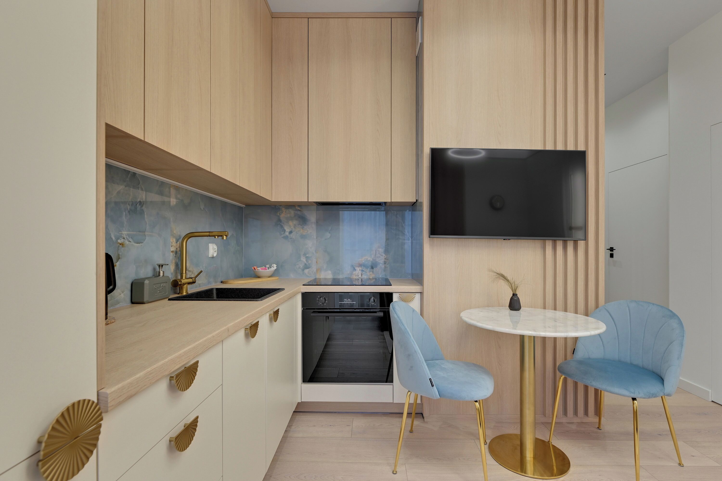 Une cuisine moderne avec des meubles en bois, un four encastré, un évier avec un robinet doré et une table ronde accompagnée de deux chaises bleues.